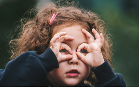 Meisje met krullen en speldje in haar haar tuurt door handen met open mond