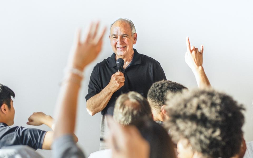 Spreker met microfoon voor publiek in aula met handen omhoog