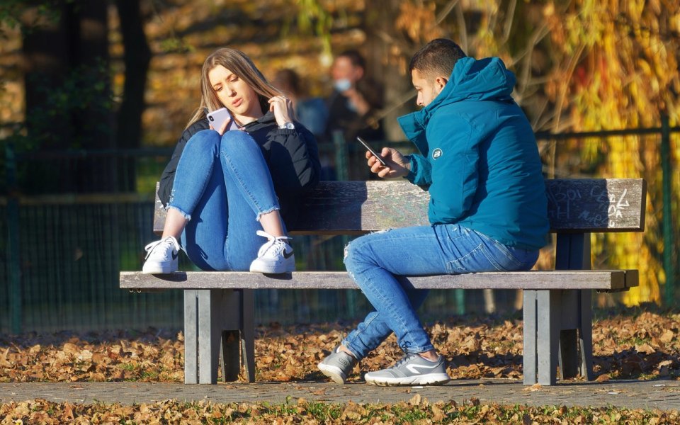 2 jongeren met gsm op een bankje buiten in de herfst