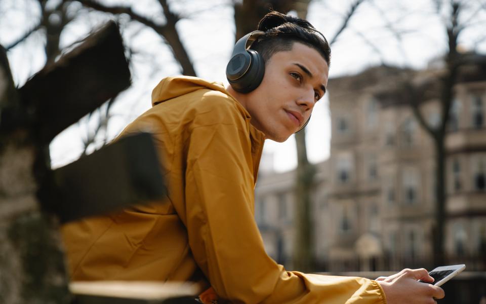 Jongere met manbun zit op stadsbankje met hoofdtelefoon op en smartphone, kijkt in de verte