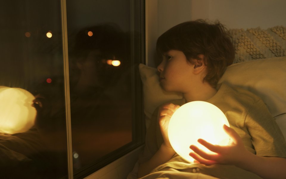 kind-in-bed--door-raam-kijkend--met-nachtlampje-in-zijn-hand