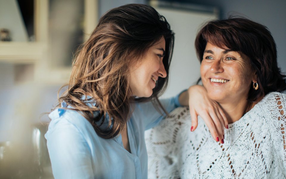 2 vrouwen, lachend, waarbij de ene haar arm op schouder van de ander vrouw legt en haar aankijkt