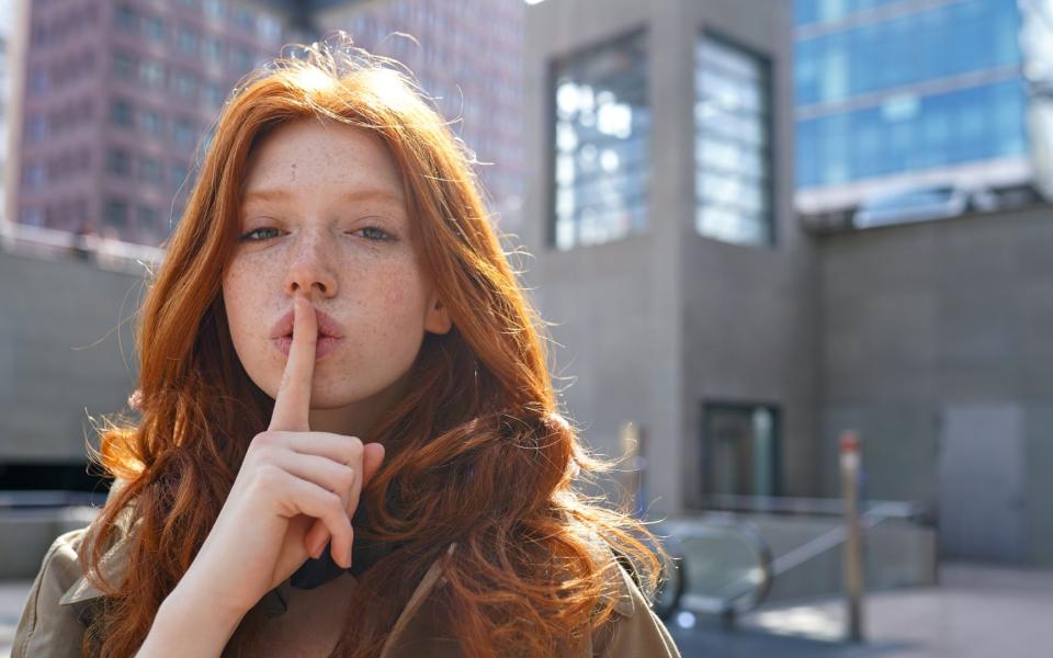 Roodharig tienermeisje in de stad met vinger op de lippen, teken van stilzwijgen