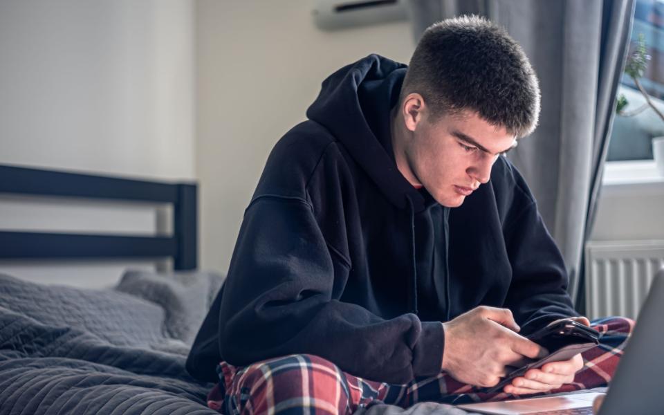 Tienerjongen zit op smartphone en laptop in zijn kamer, op bed in pyjama