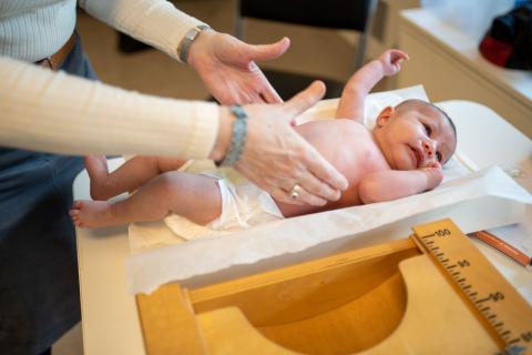 Een baby op de weegschaal in een consultatiebureau, een vrouw steekt haar handen uit naar de baby