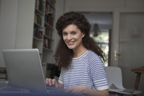 vrouw kijkt in camera met glimlach, thuiswerken aan laptop, opleiding online