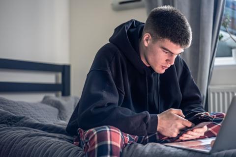 Tienerjongen zit op smartphone en laptop in zijn kamer, op bed in pyjama